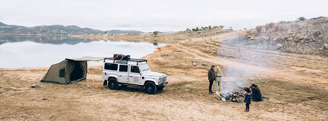 Oztent RV5 setup next to a white SUV beside a lake in a rugged outdoor setting.