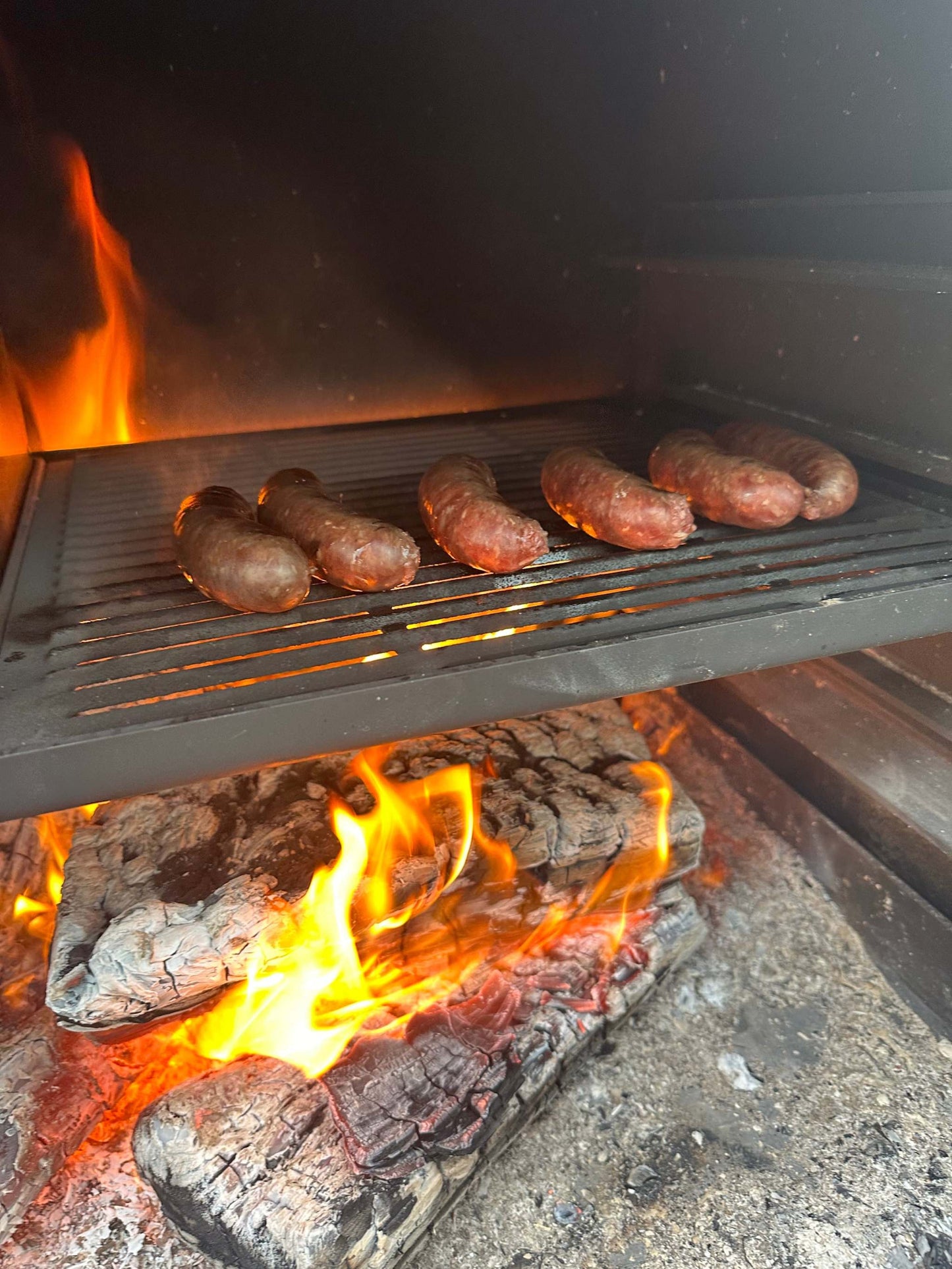 Onflame 1200 outdoor fireplace with sizzling sausages on grill over flames.