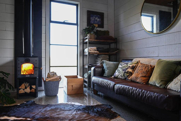 Warmington NAKED Studio 14 woodburner in a cozy room with a leather sofa and decorative cushions.